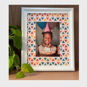 Può includere: Cornice bianca con un bordo a motivi triangolari colorati. La cornice contiene una foto di un bambino sorridente con un cappello da festa e che tiene in mano una torta di compleanno con candele accese. La cornice è su una superficie di legno.