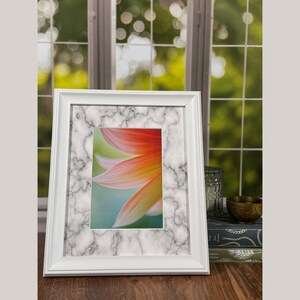 Può includere: Un quadro con cornice bianca e bordo a fantasia marmorea. L'immagine all'interno presenta un primo piano di un fiore con petali arancioni, gialli e verdi. La cornice è su una superficie di legno.