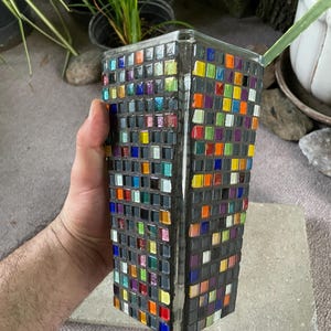 May include: A square glass vase covered in a mosaic of small, multi-colored square tiles. The tiles feature a variety of colors including red, orange, yellow, green, blue, and purple, set against a dark gray grout, creating a grid-like pattern.
