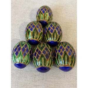 May include: Six decorative egg-shaped objects. Each egg features a blue base, green and blue diamond pattern, and gold-colored trim. The eggs are arranged in a pyramid shape on a neutral-colored surface.