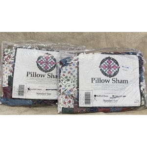 Pode incluir: Duas fronhas embaladas com estampas florais e patchwork. As fronhas são rotuladas como "Pillow Sham" e "Ruffled Sham" com um tamanho padrão de 51 x 66 cm. A embalagem é de plástico transparente.