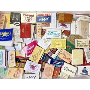 Puede incluir: Una colección de cajas de cerillas vintage con varios diseños y textos, incluyendo nombres de restaurantes y bares como "Fairbanks", "Orchards" y "Seafood Grill". Las cajas de cerillas son de varios colores y tamaños, creando una estética nostálgica.