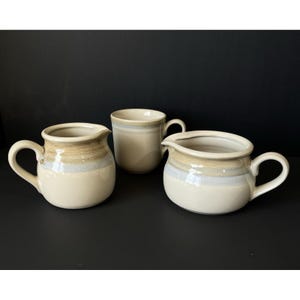 以下が含まれることがあります： クリーム、タン、ライトブルーの3色のセラミック製サービングピースのセット。セットには、ハンドル付きのピッチャー2つとマグカップが含まれています。ピッチャーには注ぎ口があります。ピースは光沢のある仕上がりです。