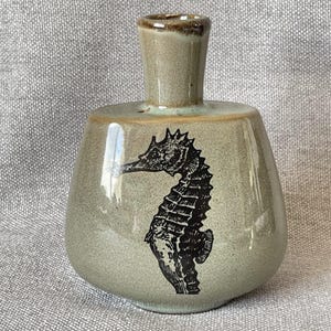 May include: A small, ceramic vase with a seahorse design. The vase is a muted green-gray color with a black seahorse illustration on the front. The top rim is a darker brown. The vase is approximately 5 inches tall.