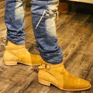 以下が含まれることがあります： マスタード色のスエードアンクルブーツ。バックル留めとローヒールが特徴です。青いデニムジーンズと合わせています。ブーツと脚の下部を写した、低いアングルからの画像です。
