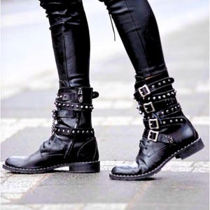 Pode incluir: Botas de couro preto com v&aacute;rias tiras, fivelas e tachas prateadas. As botas t&ecirc;m um design com atacadores e um salto baixo. As botas s&atilde;o usadas com cal&ccedil;as de couro pretas.