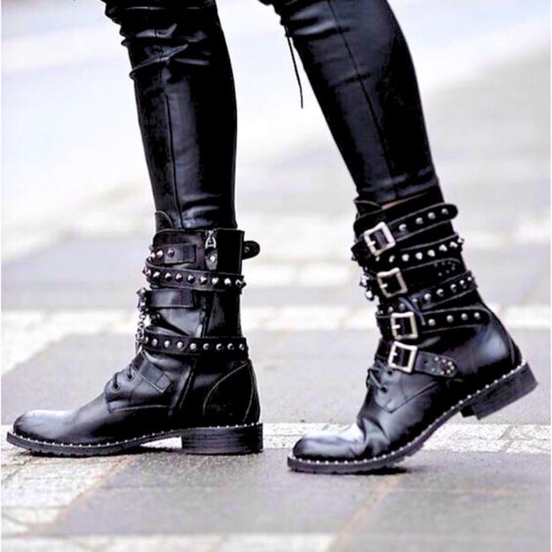 Pode incluir: Botas de couro preto com v&aacute;rias tiras, fivelas e tachas prateadas. As botas t&ecirc;m um design com atacadores e um salto baixo. As botas s&atilde;o usadas com cal&ccedil;as de couro pretas.
