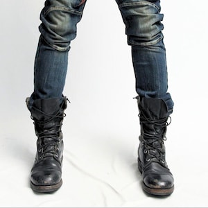 Puede incluir: Botas de combate de cuero negro con cordones, combinadas con vaqueros azules. Las botas tienen un aspecto desgastado. La imagen muestra las botas y los vaqueros desde las rodillas hacia abajo, sobre un fondo blanco.