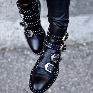 Puede incluir: Botines de cuero negro con múltiples hebillas plateadas y detalles de tachuelas. Los botines tienen punta y se usan con pantalones de cuero negro. Los botines son una declaración de moda elegante y atrevida.