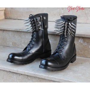 May include: Two black leather combat boots with silver metal spikes around the top. The boots feature laces, zips, and a rounded toe. The boots are displayed on a stone surface. The text "Stud Studio" is in the upper right corner.