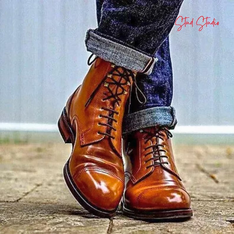 May include: A pair of polished, brown leather boots with black laces. The boots have a cap toe design and are worn with rolled-up blue denim jeans. The boots are on a wooden surface.