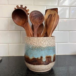 Puede incluir: Un porta utensilios de cerámica con base color crema moteada, una banda ancha de esmalte azul claro y un patrón de goteo marrón oscuro en la parte inferior. El recipiente contiene varios utensilios de cocina de madera, incluyendo un cucharón, una espumadera y una espátula.