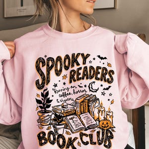 May include: A light pink crewneck sweatshirt featuring a black and orange graphic print that reads "SPOOKY READERS BOOK CLUB" and "Running on coffee, horror & chapters," with illustrations of books, a skull, candles, a pumpkin, and plants.