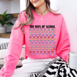 May include: A bright pink sweatshirt with the text "100 DAYS OF SCHOOL" above a grid of colorful hearts. The hearts are outlined in black and filled with various colors, including pink, yellow, blue, and purple. The sweatshirt is a crew neck style.