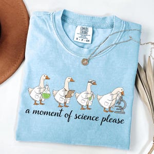 May include: A light blue t-shirt featuring a graphic of four white geese wearing lab coats and performing scientific experiments. The text "a moment of science please" is printed in black below the geese. The shirt has a Comfort Colors brand label.