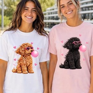 May include: A white t-shirt featuring a graphic of a brown curly-haired dog wearing pink heart-shaped sunglasses and blowing a pink bubble, and a light pink t-shirt featuring a graphic of a black curly-haired dog with pink heart-shaped sunglasses.