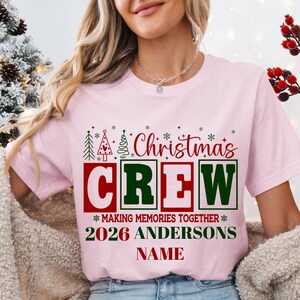 May include: A light pink t-shirt featuring a graphic with the text Christmas CREW, MAKING MEMORIES TOGETHER, 2026 ANDERSONS, and NAME. The design includes red and green block letters, stylized Christmas trees, and small snowflake icons.