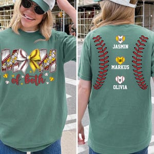 Pu&ograve; includere: Una t-shirt verde salvia con una grafica frontale che riporta il testo MOM of both e un fiocco giallo e bianco su un motivo da baseball. Il retro mostra due motivi di cuciture da baseball rosse, tre palline a forma di cuore con i numeri 12, 18, 8 e nomi.