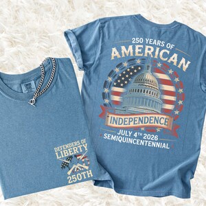 May include: Two blue t-shirts. One is folded with a silver chain necklace. The other shirt has a graphic on the back that says "250 Years of American Independence" with the Capitol building and the date July 4th 2026.