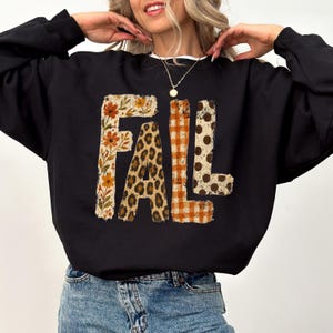 May include: A black long-sleeve sweatshirt featuring the word FALL printed in large block letters. Each letter has a distinct pattern: the F has orange flowers, the A has a leopard print, the L has orange gingham, and the L has brown polka dots.