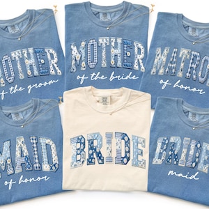 May include: A collection of blue and cream-colored t-shirts with text for a wedding party. The blue shirts have text that reads "MOTHER of the groom", "MOTHER of the bride", "MATRON of honor", "MAID of honor", and "BRIDES maid". The cream shirt says "BRIDE".