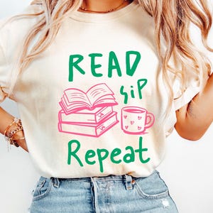 May include: A cream-coloured t-shirt featuring the text "Read Sip Repeat" in green and pink. The design includes a stack of books, an open book, and a mug with hearts. The shirt is a casual, comfortable style.