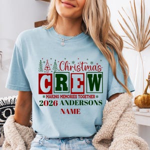 May include: A light blue short-sleeve t-shirt featuring a graphic with the text Christmas CREW MAKING MEMORIES TOGETHER 2026 ANDERSONS NAME, decorated with red and green Christmas tree icons and small snowflakes in a festive holiday design.