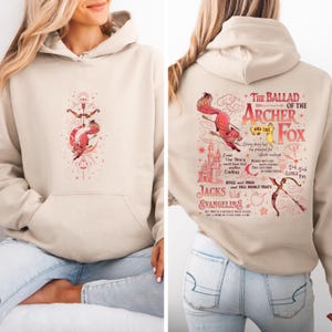 May include: Beige hoodie with a front pocket and a hood. The front features a red and gold illustration of an archer and a fox with a bow and arrow. The back displays text and illustrations related to "The Ballad of the Archer and the Fox."