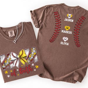 Pu&ograve; includere: Una t-shirt marrone con la scritta MOM of BOTH e un fiocco giallo e bianco a tema softball. Il retro presenta due cuciture rosse verticali da softball, tre icone a forma di cuore con i numeri 12, 18, 8 e i nomi JASMIN, MARKUS e OLIVIA.