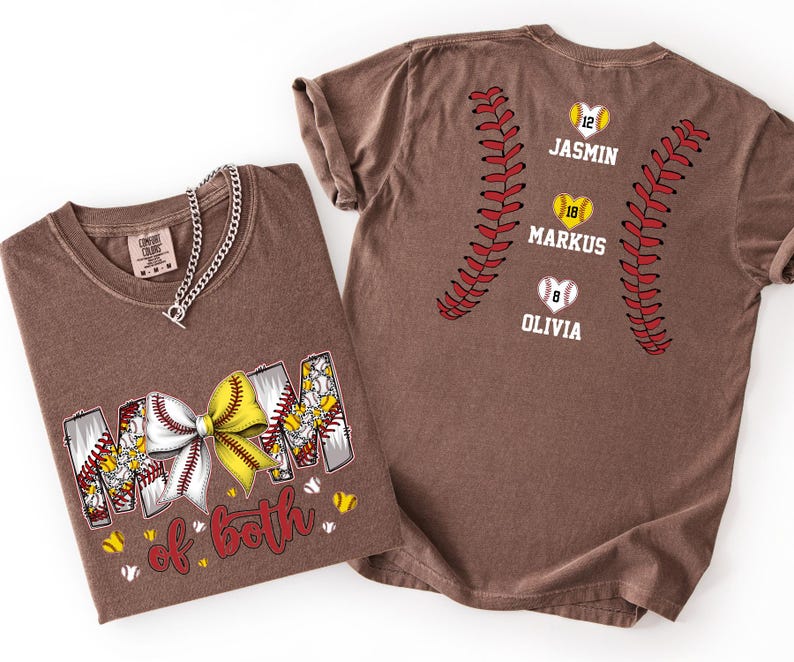 Pu&ograve; includere: Una t-shirt marrone con la scritta MOM of BOTH e un fiocco giallo e bianco a tema softball. Il retro presenta due cuciture rosse verticali da softball, tre icone a forma di cuore con i numeri 12, 18, 8 e i nomi JASMIN, MARKUS e OLIVIA.