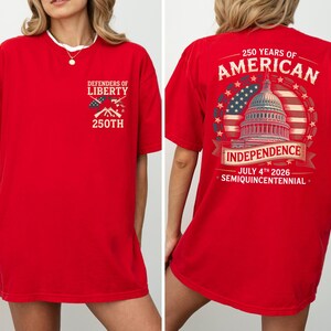 May include: Red t-shirt with patriotic graphics. The front has "Defenders of Liberty" with crossed rifles and "250th". The back shows "250 Years of American Independence" with the Capitol building, American flag, and the date "July 4th 2026".