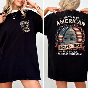 May include: Black t-shirt with a white collar. The front has the text "Defenders of Liberty" and "250th" with an emblem. The back features a design celebrating 250 years of American Independence, including the Capitol building and the date July 4th, 2026.