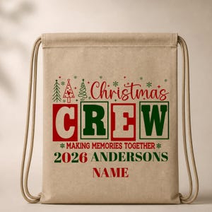 May include: A tan drawstring backpack featuring red and green text that reads Christmas CREW MAKING MEMORIES TOGETHER 2026 ANDERSONS NAME. The design includes small green and red Christmas tree icons and snowflakes above the main text.