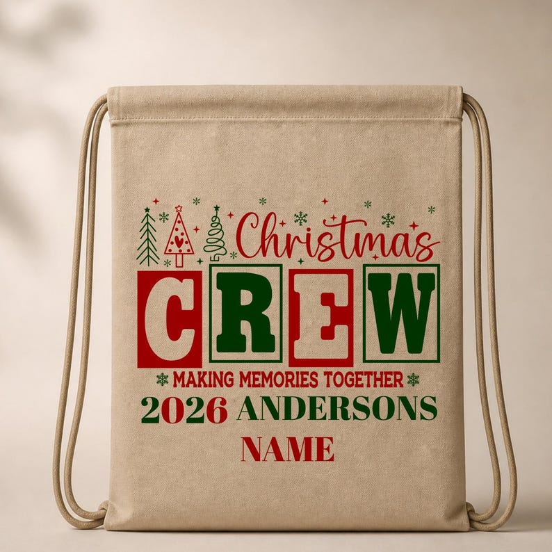 May include: A tan drawstring backpack featuring red and green text that reads Christmas CREW MAKING MEMORIES TOGETHER 2026 ANDERSONS NAME. The design includes small green and red Christmas tree icons and snowflakes above the main text.