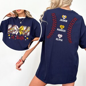Pu&ograve; includere: Una t-shirt blu navy con una grafica frontale raffigurante un fiocco da baseball e softball e il testo "MOM of both". Il retro mostra un motivo a cuciture da baseball con tre palline a forma di cuore etichettate con i nomi "JASMIN", "MARKUS" e "OLIVIA".