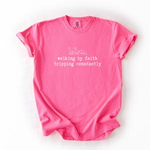 May include: A bright pink t-shirt with rolled-up sleeves. The shirt features white text and a small illustration of geese. The text reads "walking by faith tripping constantly."
