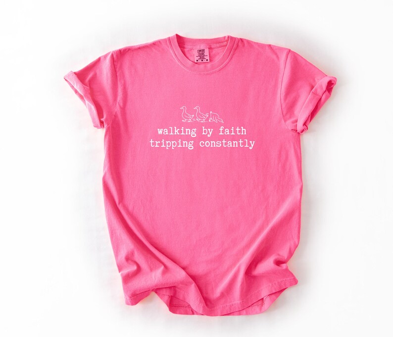 May include: A bright pink t-shirt with rolled-up sleeves. The shirt features white text and a small illustration of geese. The text reads "walking by faith tripping constantly."