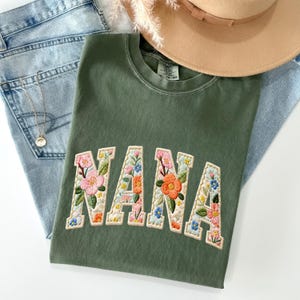 Può includere: Una maglietta verde oliva con la parola "NANA" ricamata in un disegno floreale. Le lettere sono bordate di color crema e riempite di fiori rosa, arancioni e blu. La maglietta è abbinata a jeans blu e un cappello beige.
