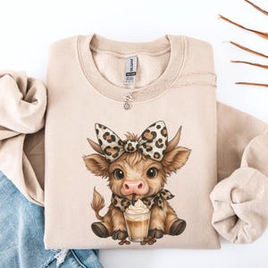 May include: A beige sweatshirt featuring a cartoon cow with a leopard print bow, scarf, and a coffee drink. The cow is brown and tan with large eyes. A silver necklace is draped across the sweatshirt.