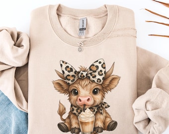 Puede incluir: Sudadera beige con un diseño de vaca de dibujos animados. La vaca tiene un lazo y una bufanda con estampado de leopardo y está bebiendo café. Un collar plateado está drapeado sobre la sudadera. La vaca es marrón y beige.
