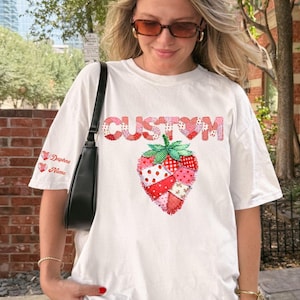Puede incluir: Camiseta blanca con un diseño de fresa de patchwork y la palabra "CUSTOM" encima. La manga izquierda tiene el texto "Daphne Name" con gráficos de corazón. El diseño utiliza colores rojo, rosa y verde.