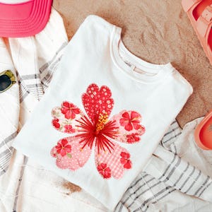 Könnte beinhalten: Weißes T-Shirt mit einem großen, farbenfrohen Blumendesign. Die Blume hat rote und rosa Blütenblätter mit einem detaillierten Zentrum. Das T-Shirt liegt auf einem gestreiften Handtuch, mit einer rosa Kappe und Sonnenbrille.