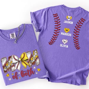 Pu&ograve; includere: Una t-shirt viola con una grafica sul davanti che recita MOM of both con un fiocco da softball giallo e bianco. Il retro della maglietta mostra due lacci da softball rossi, tre softball gialli a forma di cuore con i numeri 12, 18, 8 e i nomi JASMIN, MARKUS, OLIVIA.