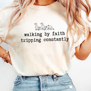 May include: Cream-colored t-shirt with the text "walking by faith tripping constantly" in black, along with a line drawing of three ducks. The shirt is made of a soft material and has a relaxed fit.