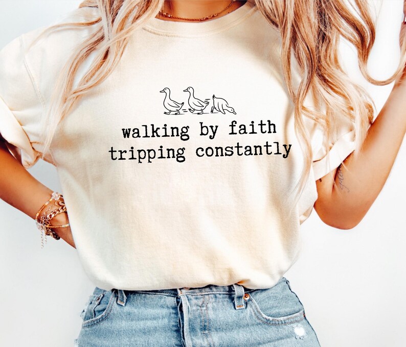 May include: Cream-colored t-shirt with the text "walking by faith tripping constantly" in black, along with a line drawing of three ducks. The shirt is made of a soft material and has a relaxed fit.