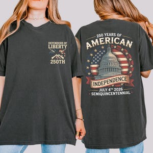 May include: Dark grey t-shirt with "Defenders of Liberty" and "250th" printed on the front. The back features a design commemorating 250 years of American Independence, including the Capitol building, the American flag, and the date July 4th, 2026.