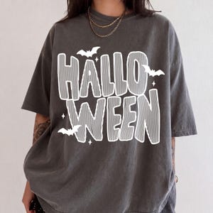 May include: A charcoal gray short-sleeve t-shirt featuring the white text HALLOWEEN in a bold, striped font. The design is accented with small white silhouettes of flying bats and four-pointed stars scattered around the lettering.