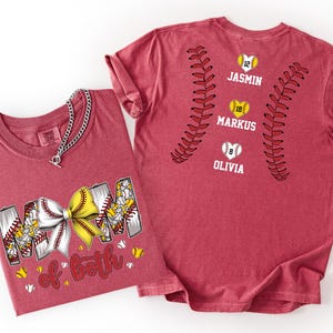 Pu&ograve; includere: Una t-shirt rossa con una grafica che riporta il testo "MOM of both" e un fiocco giallo e bianco. Il retro della maglietta mostra due motivi curvi di cuciture da baseball, tre icone di baseball a forma di cuore con i numeri 12, 18 e 8, e i nomi "JASMIN", "MARKUS" e "OLIVIA".