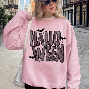 May include: A light pink long-sleeve crewneck sweatshirt featuring the word HALLOWEEN printed in large black letters with a vertical striped pattern. The design includes small black bat silhouettes and four-pointed star accents scattered around the text.