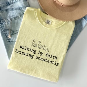 May include: A light yellow t-shirt with the text "walking by faith tripping constantly" and a graphic of three ducks. The shirt is folded on top of a pair of light blue jeans and a tan hat.